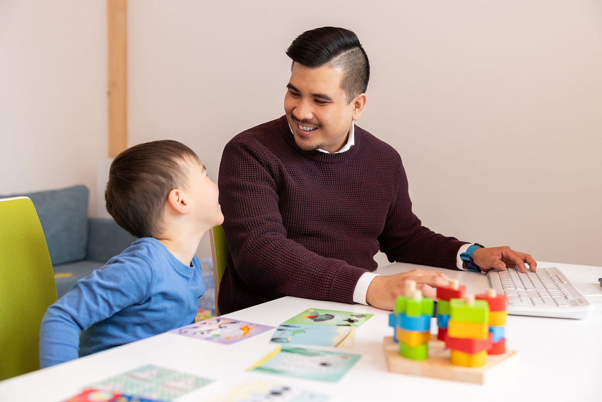 Vereinbarkeit von Familie und Beruf. (Foto: UW/H | Michael Schwettmann) Ein Vater sitzt gemeinsam mit seinem Sohn im Eltern-Kind-Raum der Uni. Vor ihnen auf dem Tisch liegen neben der Tastatur des Arbeitsrechners verschiedene Spielsachen ausgebreitet.