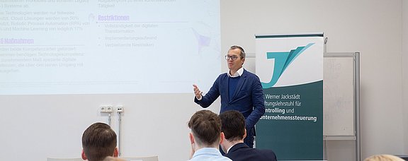 Wir bringen Praxis in den Seminarraum (Foto: UW/H | Miriam Kreimeyer) Ein Mann im Anzug steht in einem Seminarraum und spricht zu Studierenden. Hinter ihm an der Wand wird eine Präsentation gezeigt.