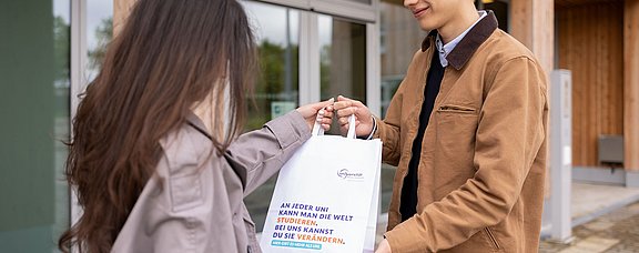 Erlebe den Unialltag an der UW/H als Student:in für einen Tag. (Foto: UW/H | Michael Schwettmann) Junger Mann überreicht einer jungen Frau eine Papiertüte.