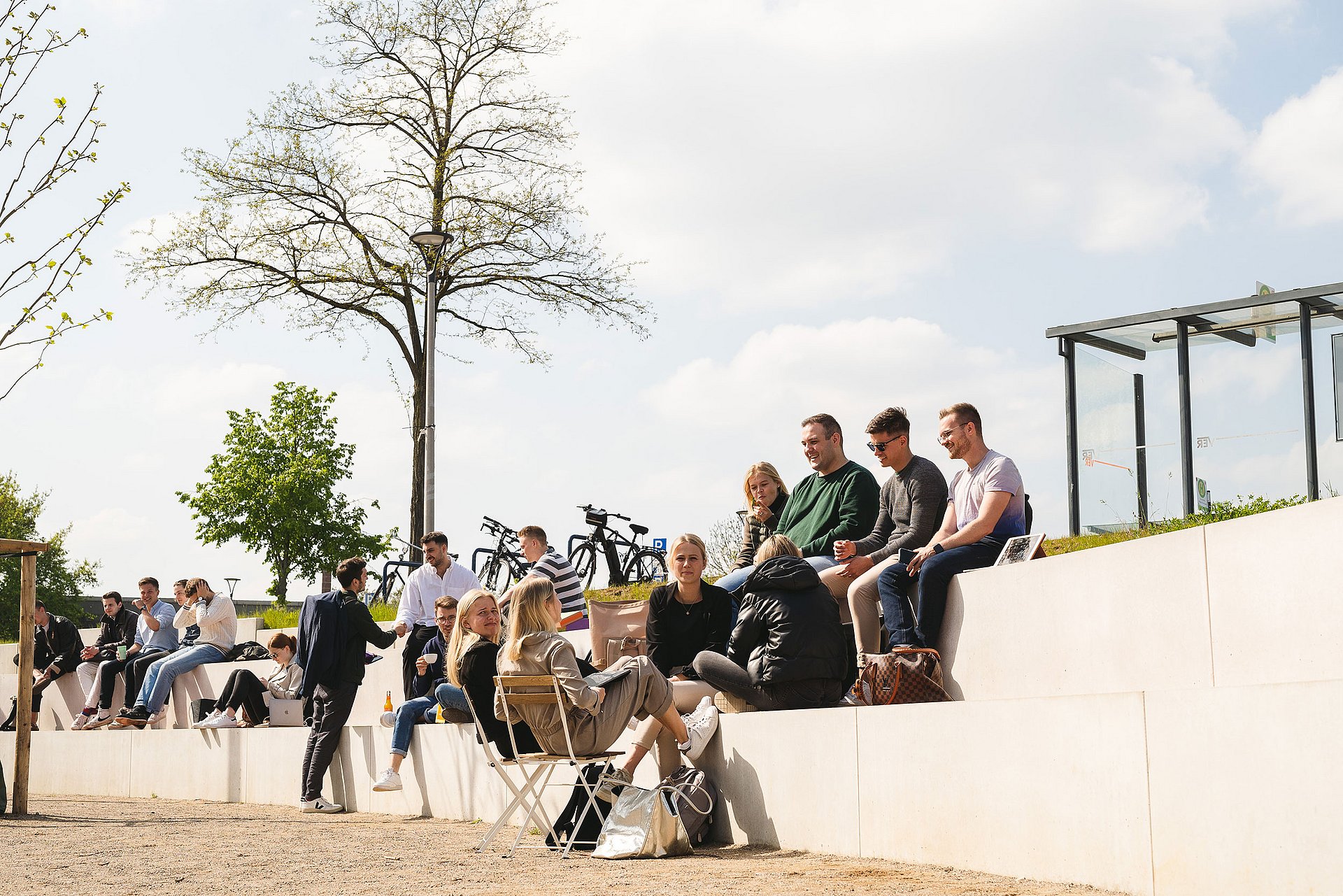 Der Campusplatz vor dem Holzgebäude – beliebter Treffpunkt & Aufenthaltsort (Foto: UW/H | Johannes Buldmann) Studierende sitzen auf Treppenstufen draußen auf dem Vorplatz der Universität Witten/Herdecke