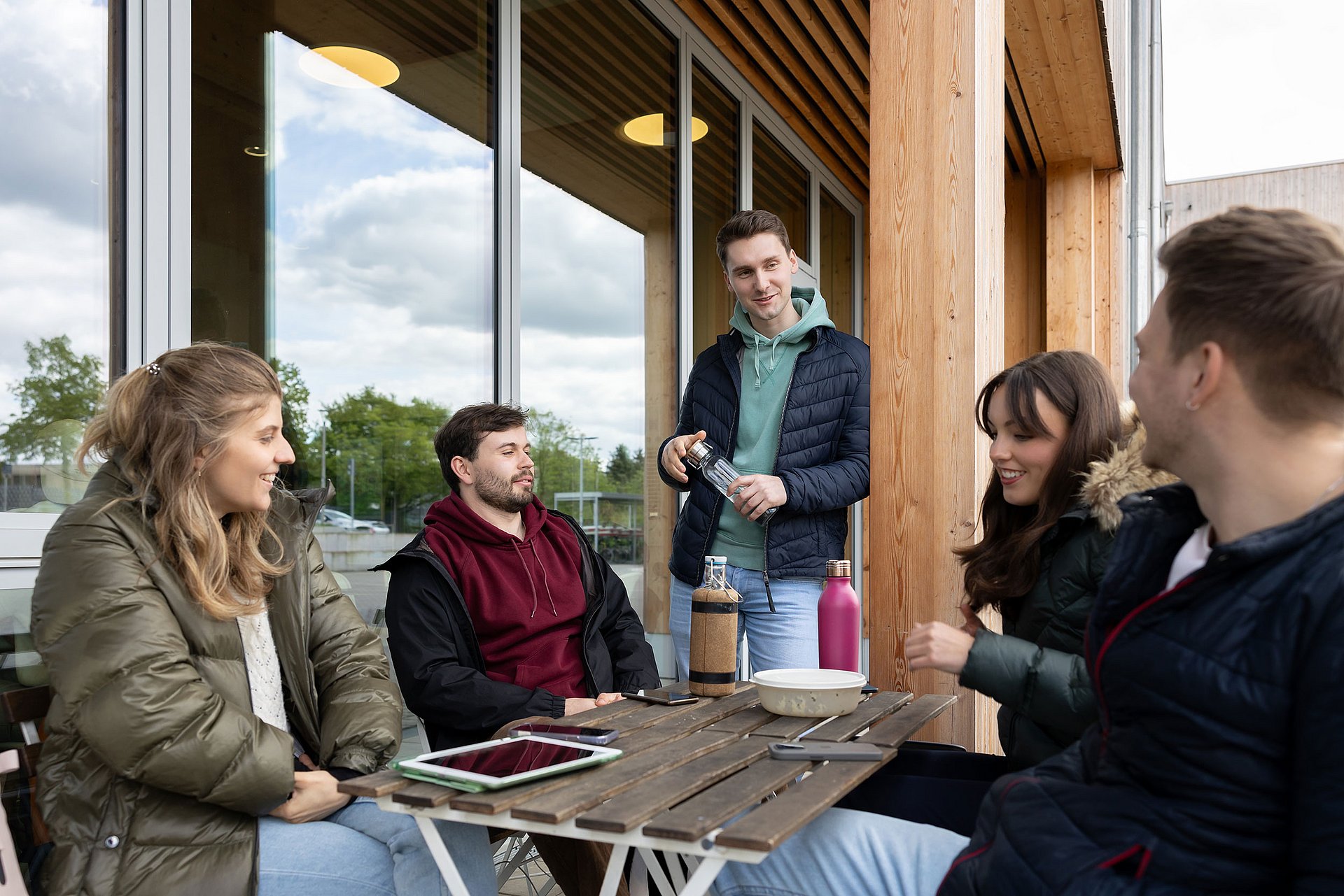 Entspannte Pausenzeit zwischen den Seminaren (Foto: UW/H | Michael Schwettmann) Eine Gruppe Studierender sitzt an einer Terrassen-Sitzgelegenheit vor dem Uni-Café und unterhält sich.